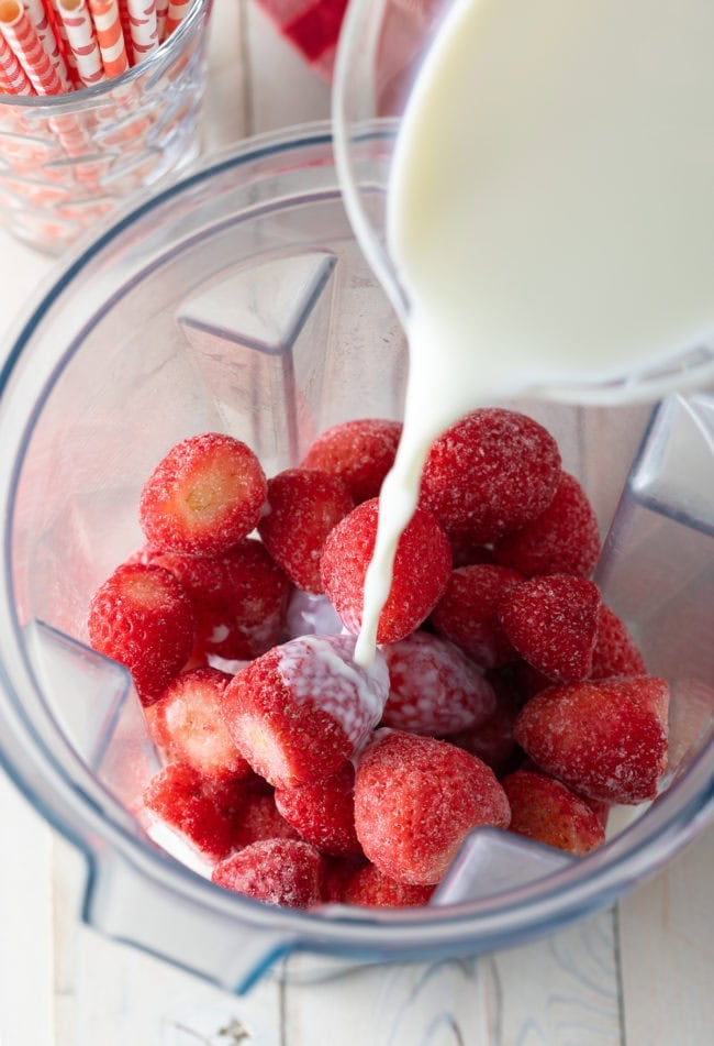 Frozen strawberries in a blender with milk being poured on top.