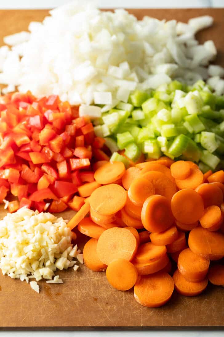 Chopped fresh ingredients in piles on a wood cutting board: Onion, red peppers, carrots, garlic, and celery.