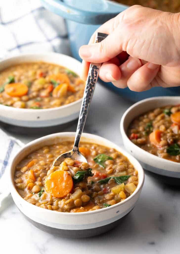 Hand with a spoon scooping a serving of traditional lentil soup from a white bowl.