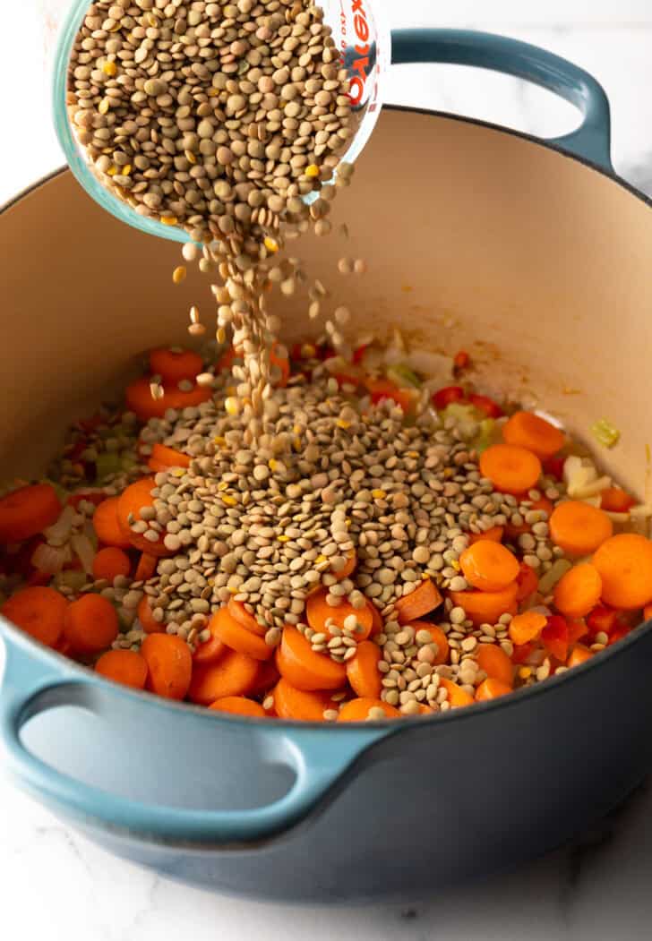 Adding dried lentils from a measuring glass to a cooking pot with carrots.